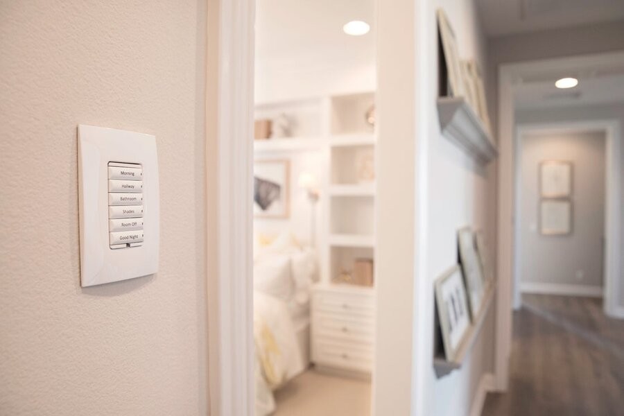A well-lit hallway with a door leading into an illuminated room, with a Control4 wall panel in the hallway displaying lighting control system buttons.